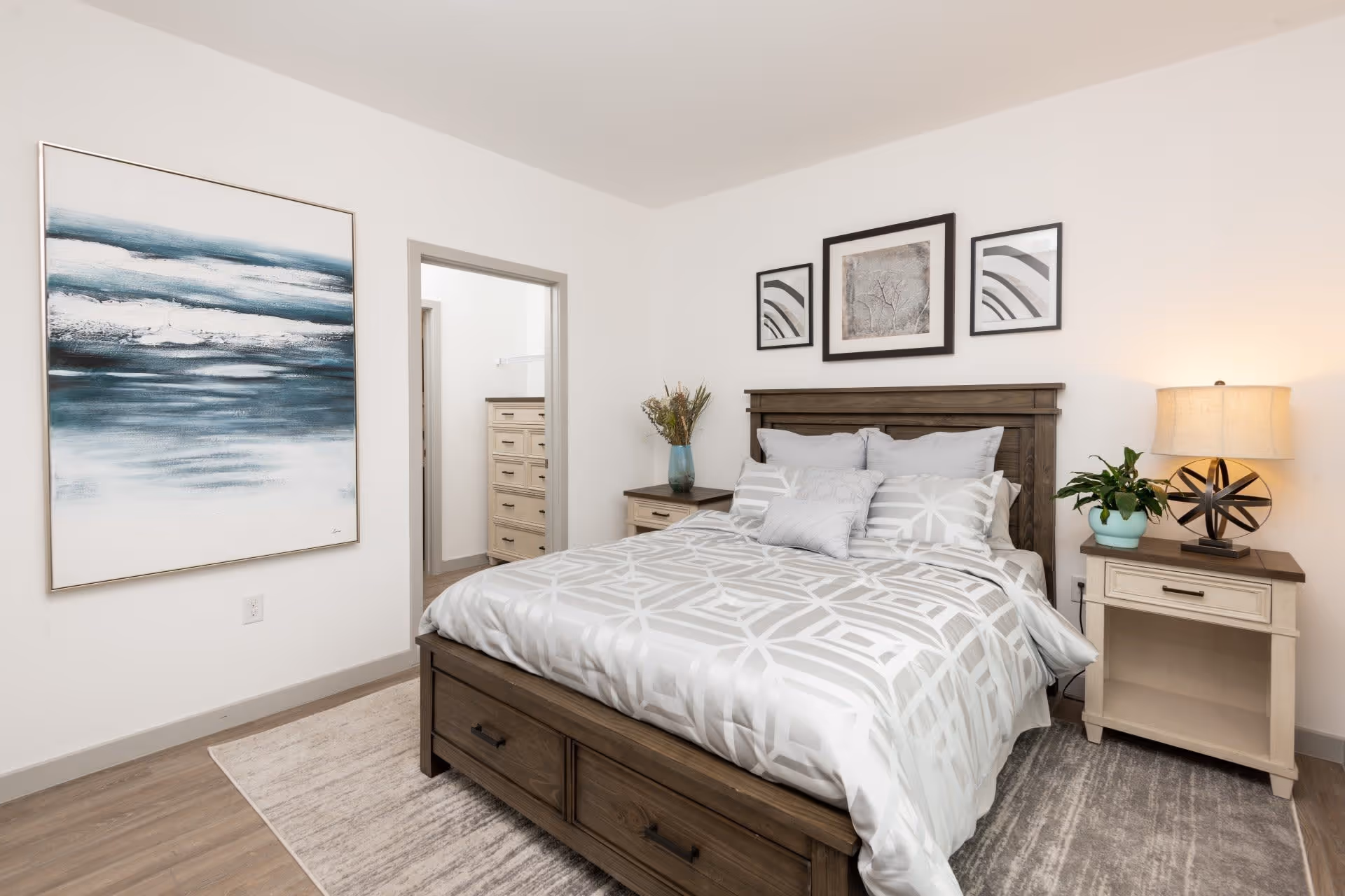 A neatly made bed with a wooden headboard and white and gray patterned bedding in a bedroom. There are three framed artworks on the wall above the bed. On each side of the bed are nightstands; the left one has a vase with dried flowers, and the right one has a lamp and a small potted plant. A large abstract painting with blue and white tones hangs on the adjacent wall. The floor has a light-colored rug and wood flooring. An open doorway reveals a glimpse of a dresser in an adjoining room.