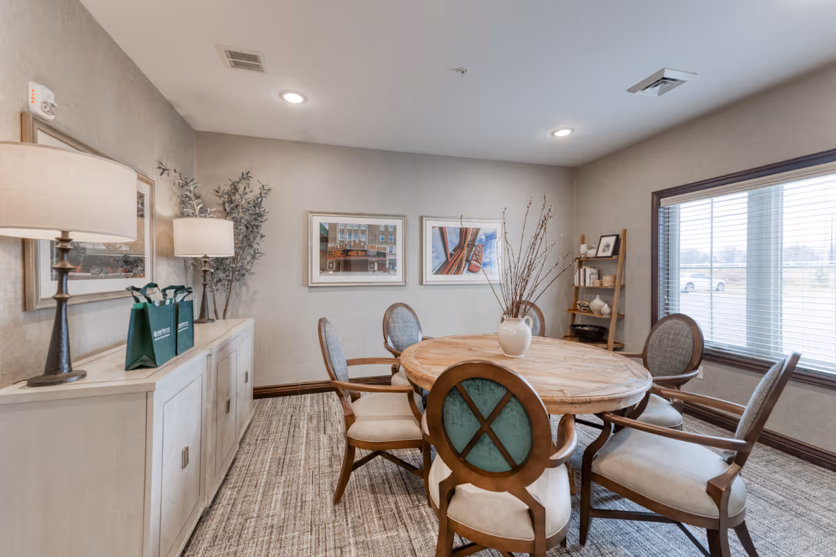 A cozy dining room in a senior living facility featuring a round wooden table surrounded by six cushioned chairs. The room has beige walls with framed artwork, a large window with blinds letting in natural light, a sideboard with two table lamps and green gift bags, and a small wooden shelf with decorative items.