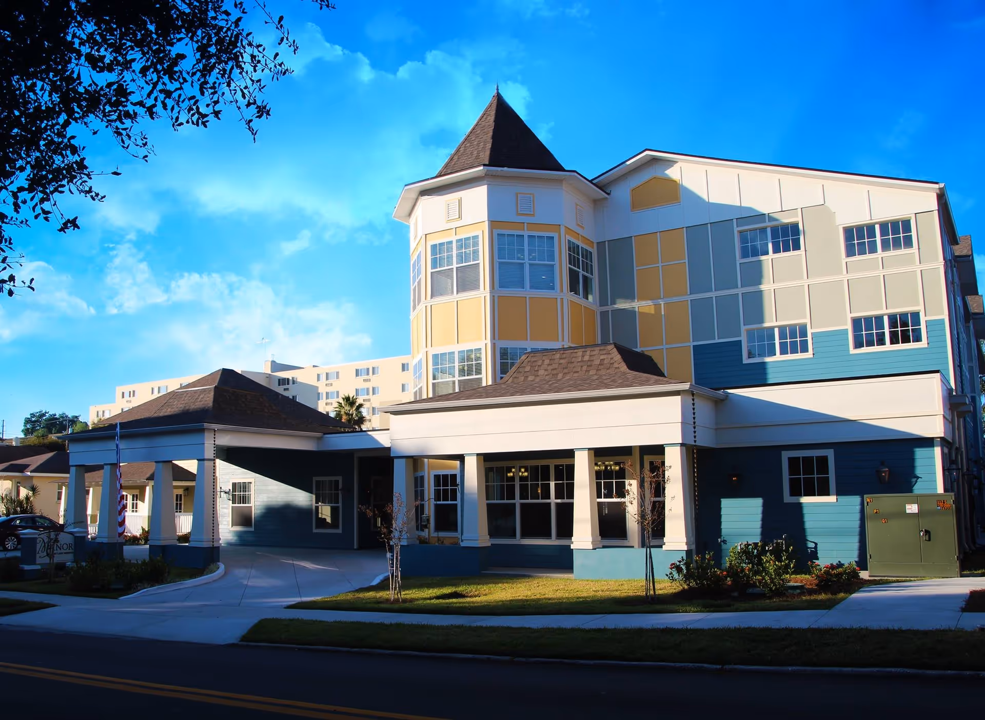 Exterior view of The Manor at Lake Morton, a multi-story senior living facility with a colorful facade featuring blue, yellow, and beige panels, a peaked tower, and a covered entrance driveway. The building is surrounded by a well-maintained lawn and small trees under a bright blue sky with some clouds.