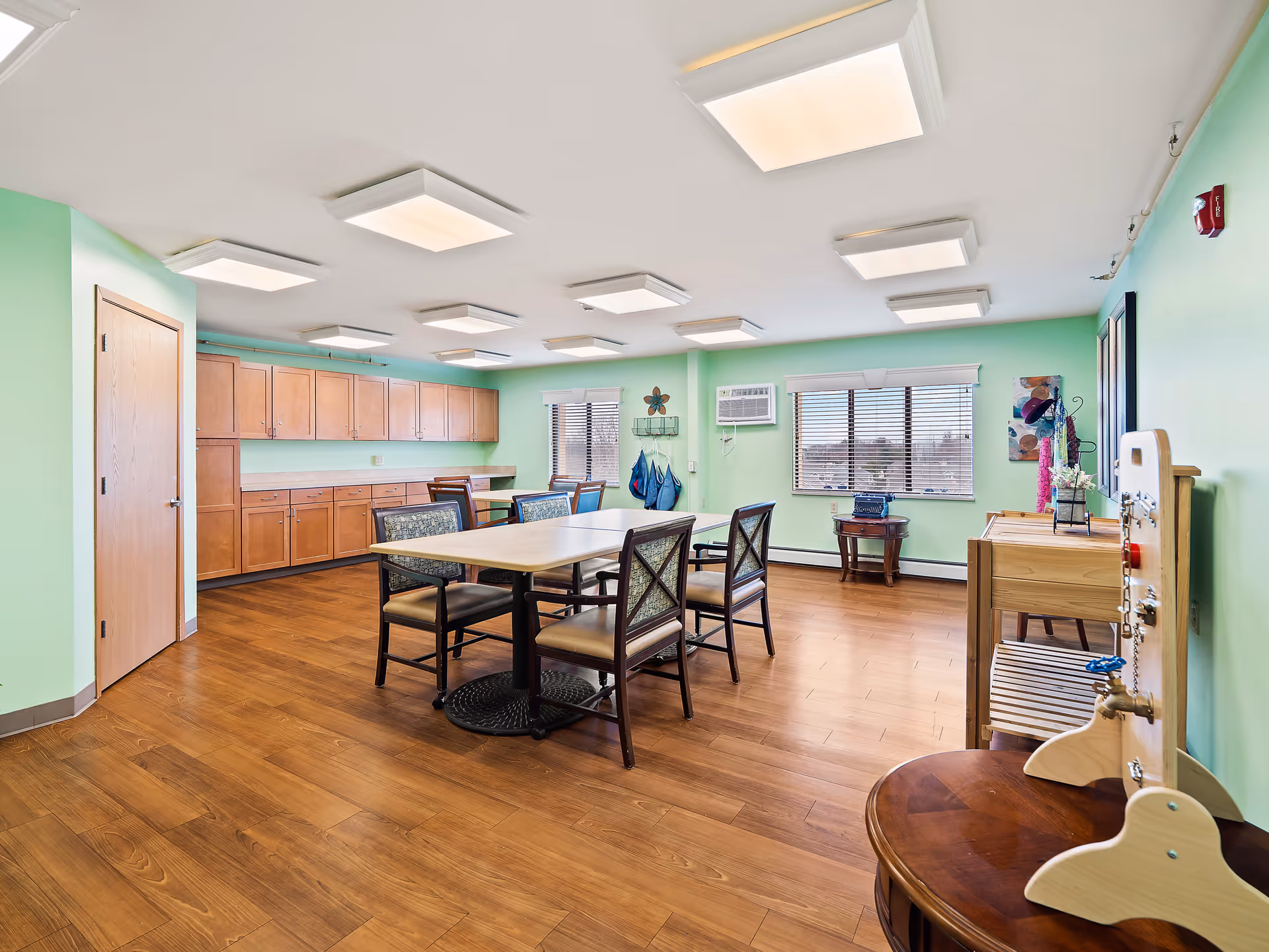A bright room with light green walls and wooden flooring featuring a rectangular table surrounded by six chairs. The room has multiple ceiling lights, wooden cabinets along one wall, two windows with blinds, and various small furniture pieces and decorations.