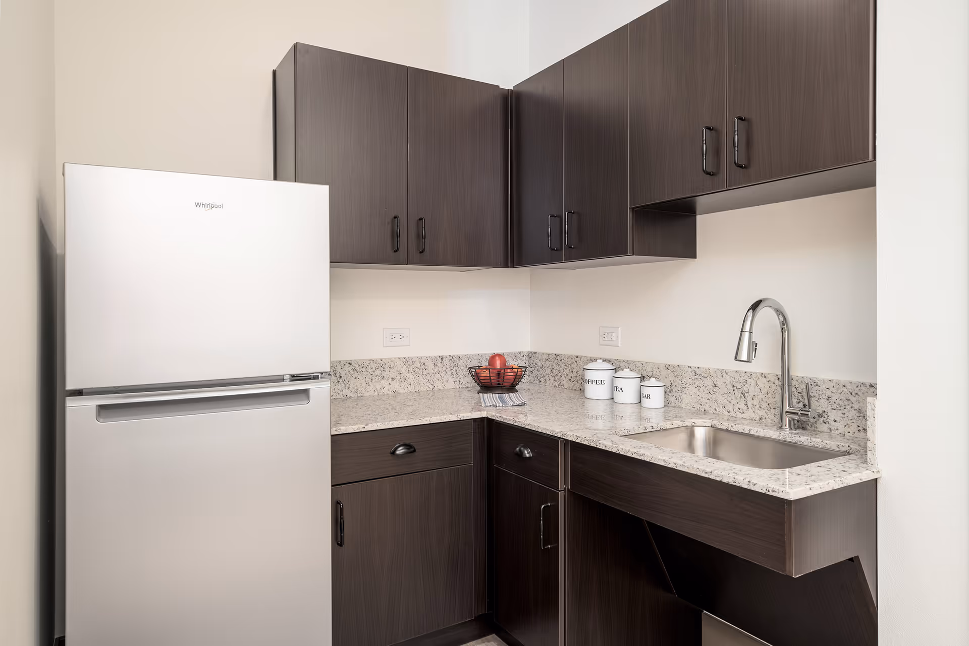 A modern kitchen corner with dark wood cabinets, a stainless steel Whirlpool refrigerator, a granite countertop with a built-in sink and chrome faucet. On the countertop, there is a black wire fruit basket with red apples and three white canisters labeled Coffee, Tea, and Sugar.