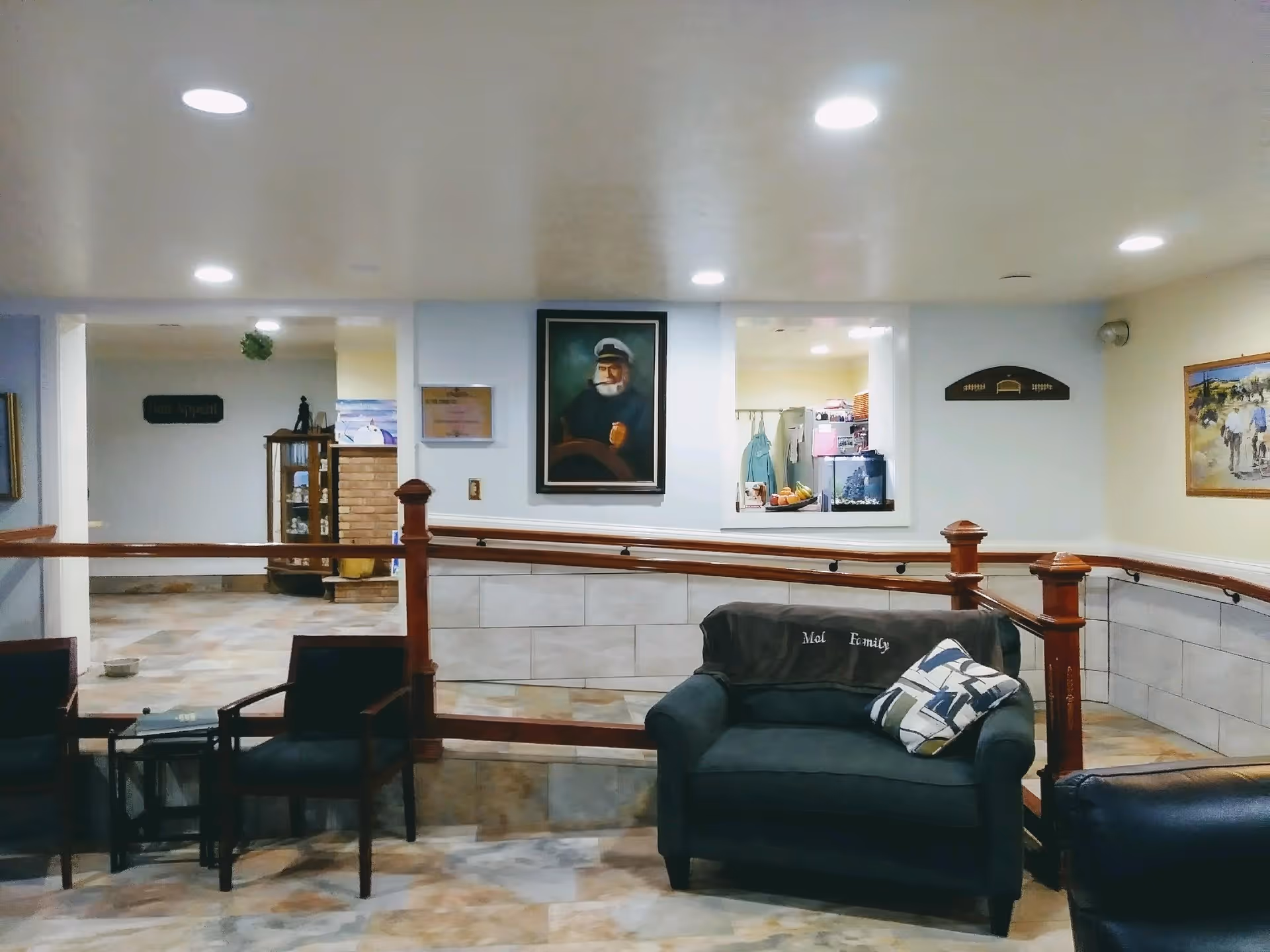 Interior view of a senior living facility common area with a dark armchair and two chairs around a small table. The area has tiled flooring, wooden railing, and several paintings on the walls, including a portrait of a man in a captain's hat. There is a window opening to another room with visible kitchen items.