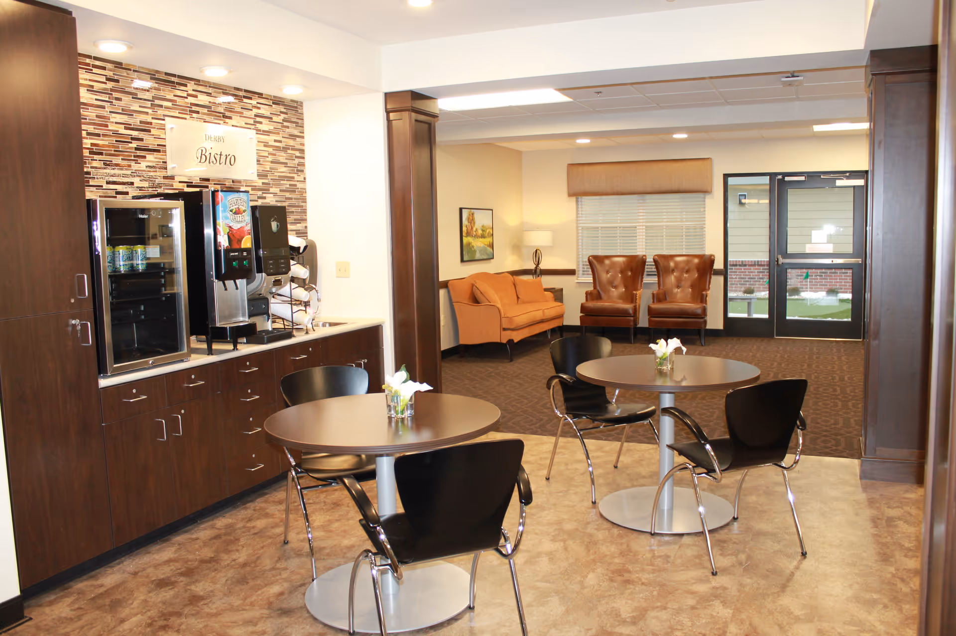 A bistro-style common room with round tables and black chairs, a coffee station and cabinets to the left, and lounge seating in the background.
