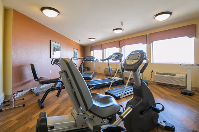 A small fitness room with exercise equipment including a recumbent bike, treadmill, and rowing machine. The room has wood flooring, beige and orange walls, three ceiling lights, and large windows with blinds letting in natural light. There are framed pictures on the walls and an air conditioning unit below the windows.