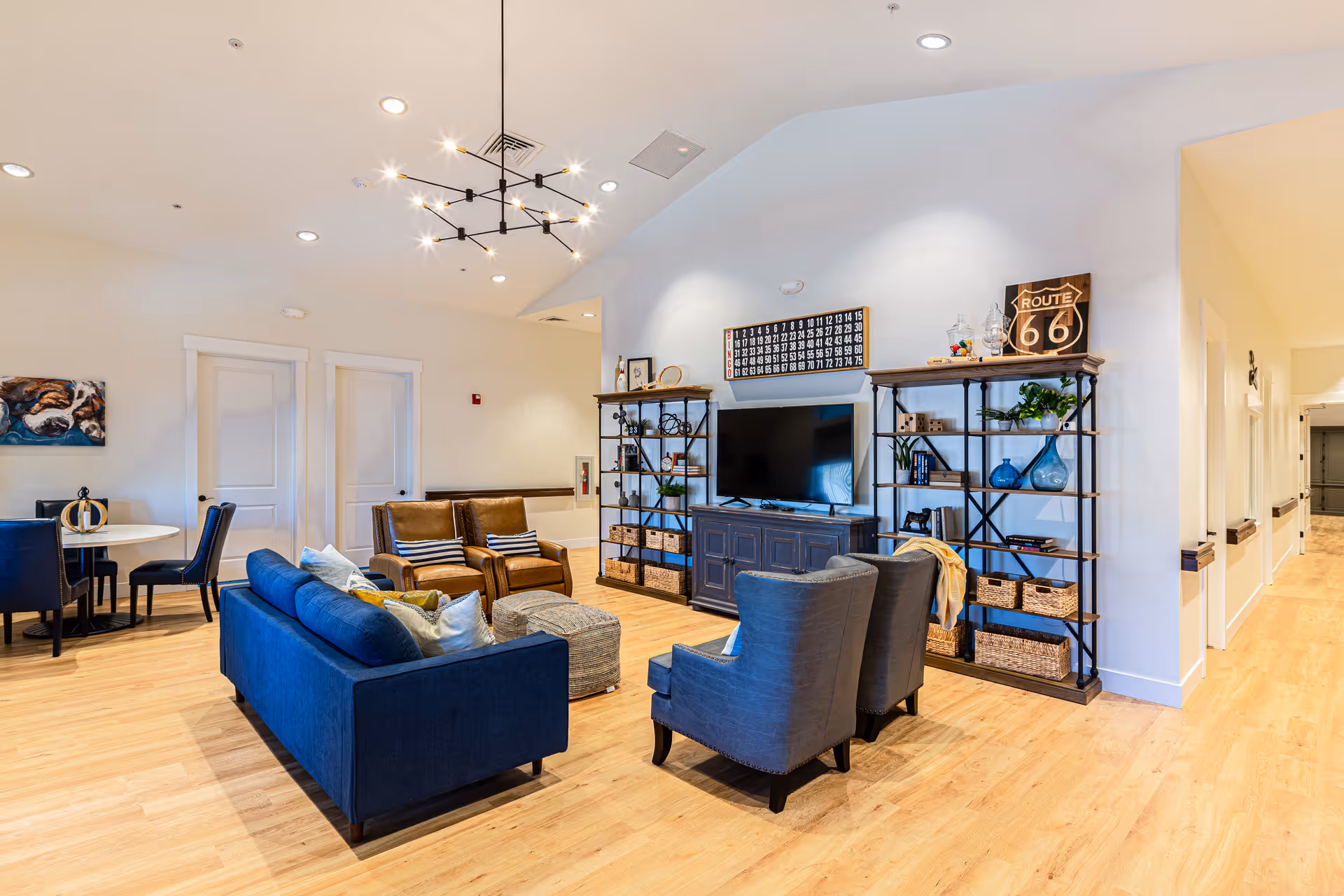A bright and spacious living room area in an assisted living facility featuring a blue sofa, two brown leather chairs, and two gray armchairs arranged around a small ottoman. There is a large flat-screen TV mounted on a dark wooden cabinet flanked by two tall shelving units with decorative items and plants. A modern chandelier hangs from the ceiling, and a small dining table with four chairs is visible in the background. The room has light wooden flooring and white walls.
