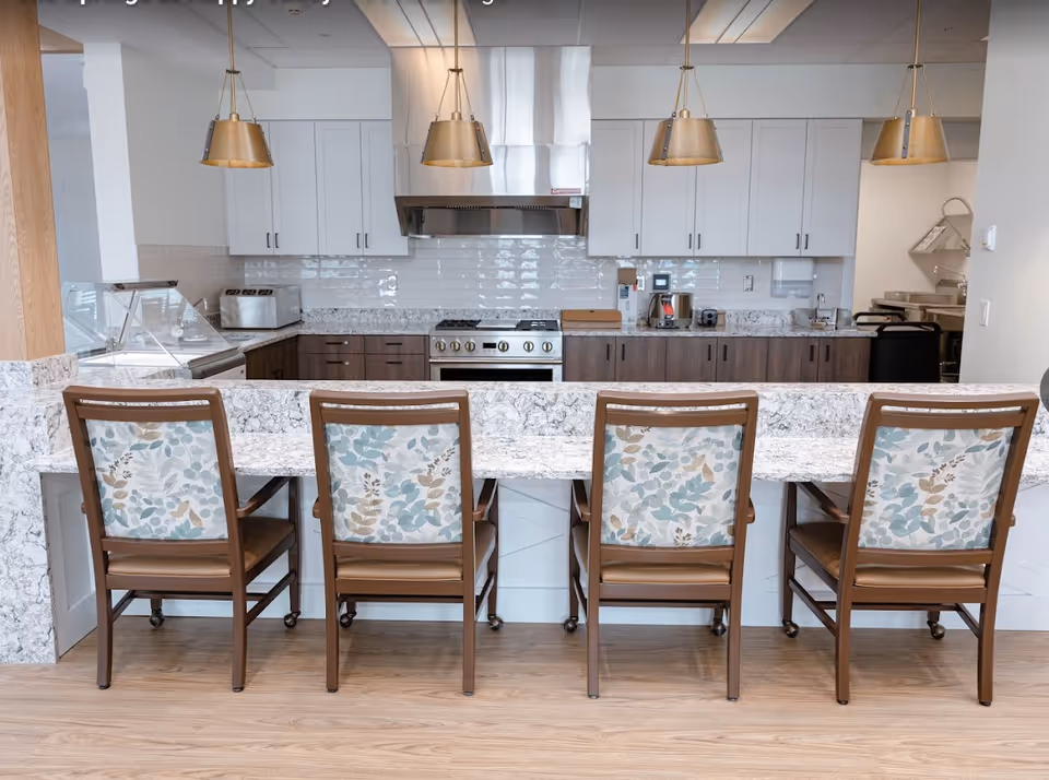 Four patterned-back chairs along a marble counter facing a modern kitchen with white cabinets, stainless range hood, and pendant lights.