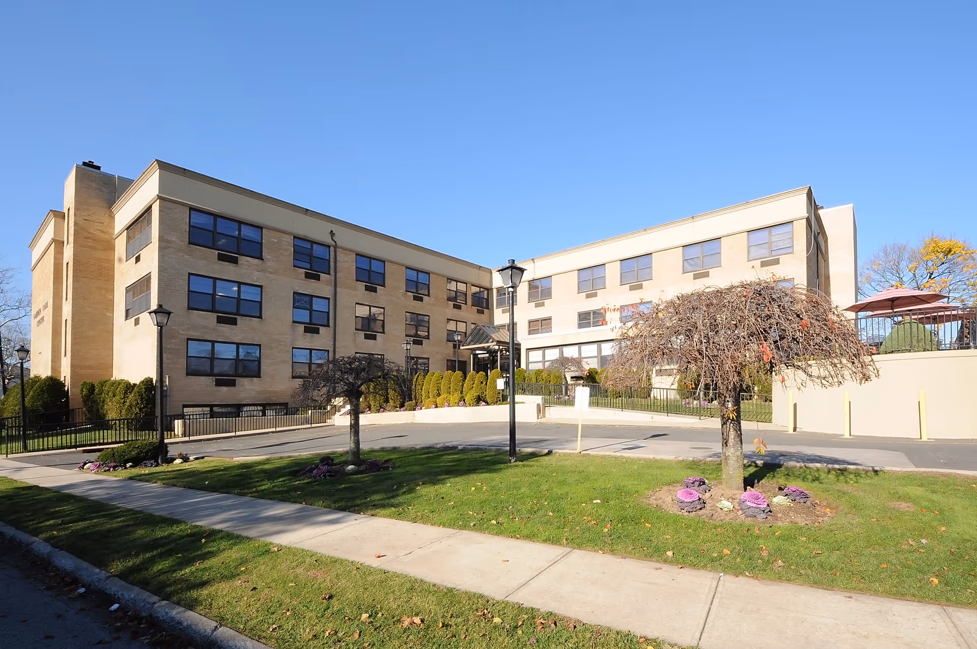 Front exterior of a three-story beige brick senior living building with small trees, lawn, and a sidewalk under a clear blue sky.