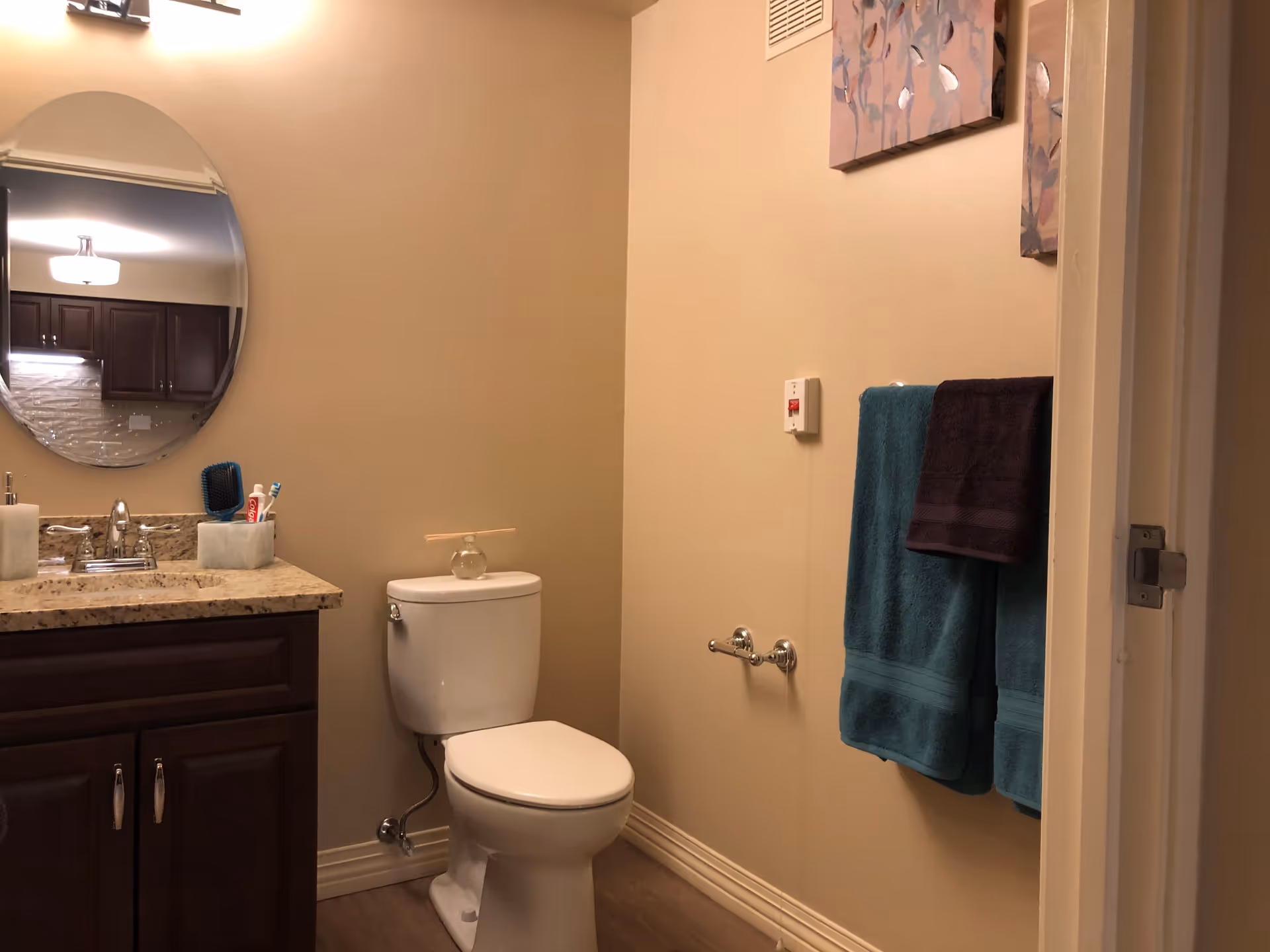 Small bathroom with a toilet, dark wood vanity with granite countertop and oval mirror, and a towel rack holding teal towels.