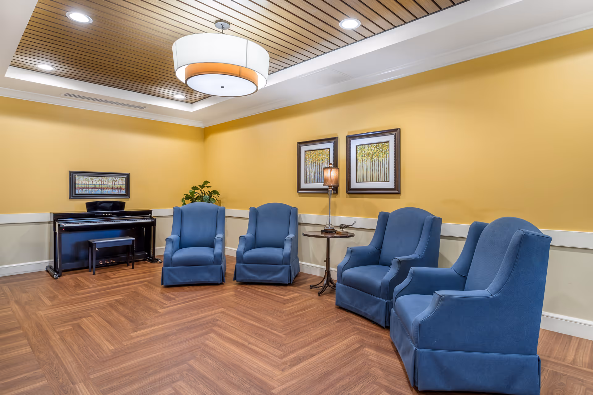 A cozy seating area with five blue upholstered armchairs arranged in a semi-circle on a wooden floor. The walls are painted yellow with white trim, and there are three framed paintings of trees hanging on the walls. A small round table with a lamp and decorative items is placed between two of the chairs. A black upright piano with a matching bench is positioned against the wall, accompanied by a potted plant. The ceiling features wooden slats and recessed lighting with a large round pendant light in the center.