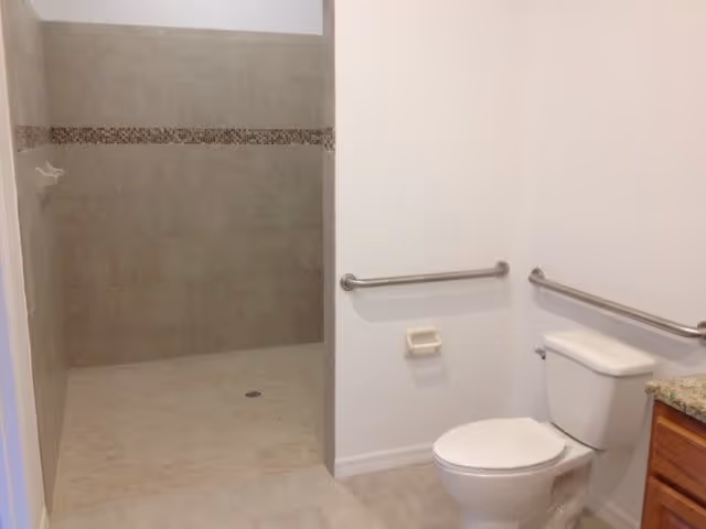 A clean and accessible bathroom featuring a white toilet with metal grab bars on the walls beside and behind it, a tiled walk-in shower with a decorative tile border, and a granite countertop partially visible on the right.