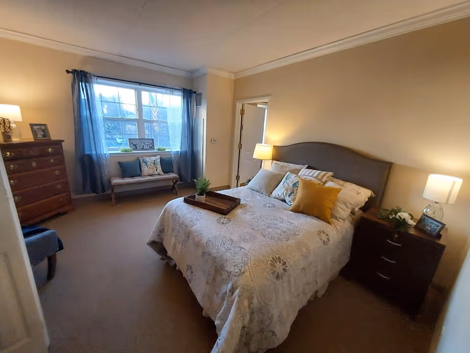 A cozy bedroom with a bed covered in a patterned quilt and multiple pillows, two bedside tables each with a lamp, a wooden dresser with a framed photo and lamp, a bench with cushions under a large window with sheer curtains, and beige walls.