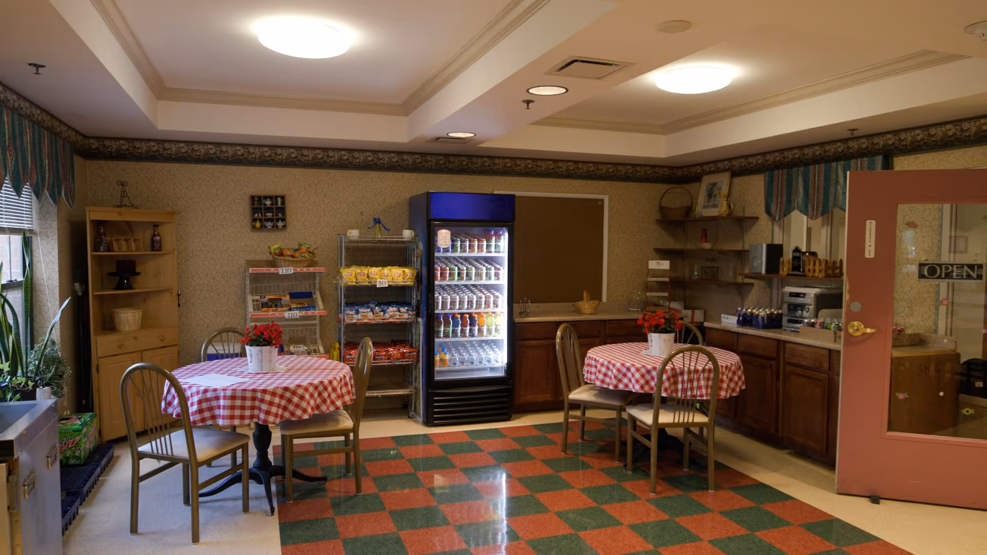 A cozy dining area with two round tables covered in red and white checkered tablecloths, each with a small potted plant centerpiece. There are metal chairs around the tables. Against the wall is a refrigerator stocked with beverages, metal racks with snacks, wooden cabinets, and shelves with various items. The floor has a red and green checkered pattern, and the walls have a floral wallpaper border near the ceiling. A door with an 'OPEN' sign is visible on the right side.