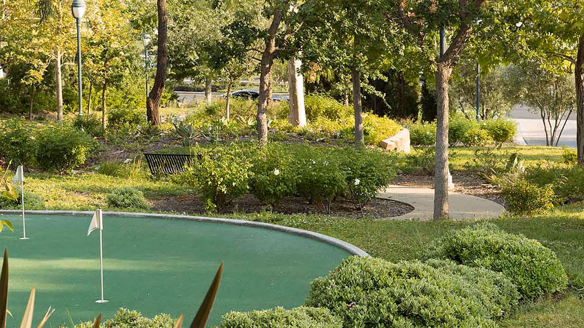 A landscaped outdoor area with a putting green featuring three small white flags, surrounded by green bushes, trees, and a curved walking path. There is a black bench partially visible among the greenery.