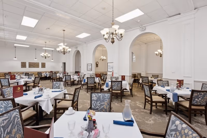 A spacious dining room with multiple tables covered in white tablecloths, each set with glassware, napkins, and menus. The room features patterned chairs, chandeliers hanging from the ceiling, and arched doorways along the walls.