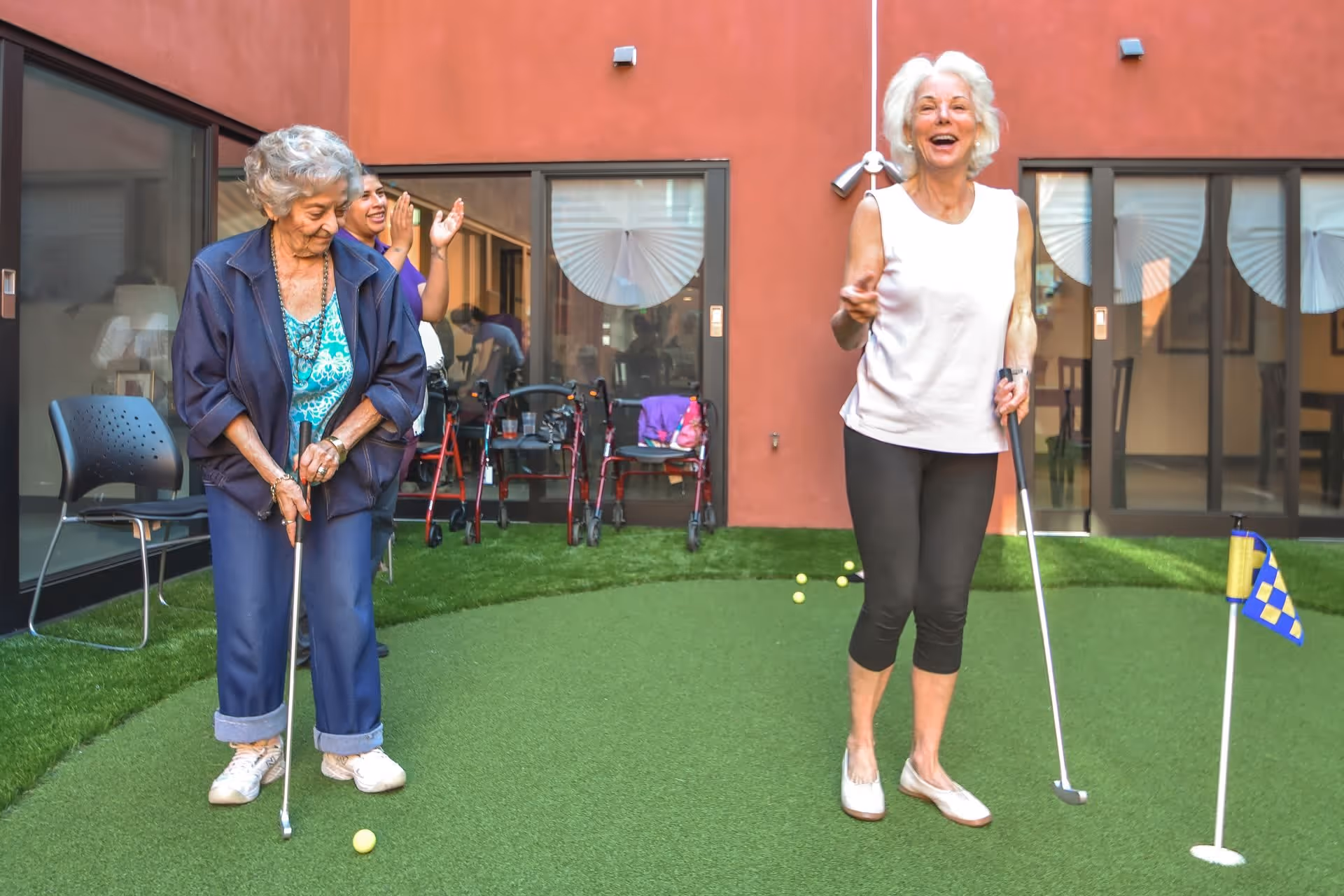 Two elderly women playing mini golf on an artificial putting green in an outdoor courtyard area of a senior living facility. One woman is preparing to putt while the other is smiling and holding a golf club near the hole with a blue and yellow checkered flag. In the background, a staff member is clapping and there are walkers and chairs near the building windows.