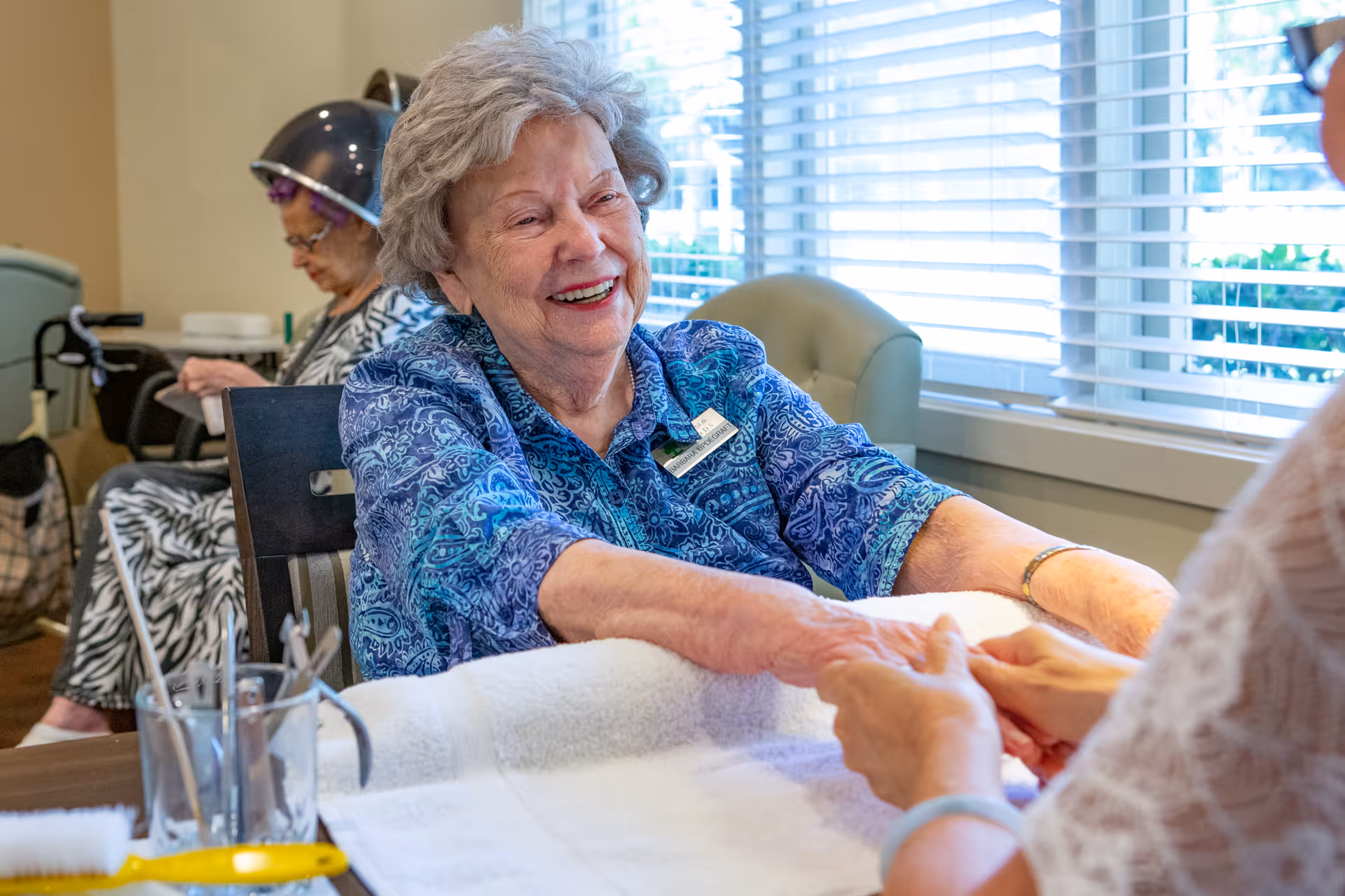 An elderly woman with gray hair and a blue patterned shirt is smiling while receiving a hand massage or manicure from another person. In the background, another elderly woman is sitting under a hair dryer in a salon setting with large windows and blinds letting in natural light.