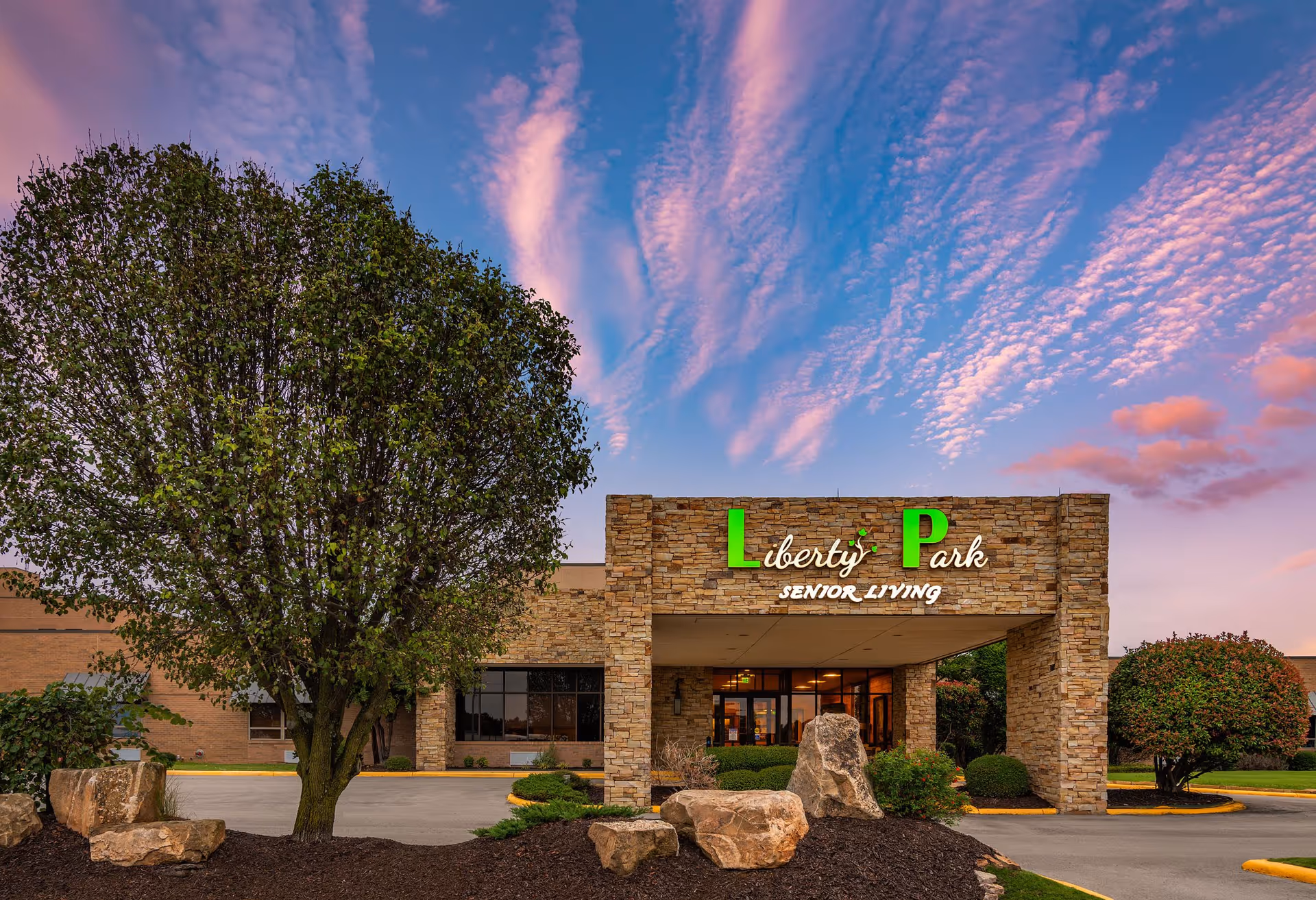 Stone-faced entrance of Liberty Park Senior Living with illuminated signage, landscaped trees, rocks, and a colorful evening sky.