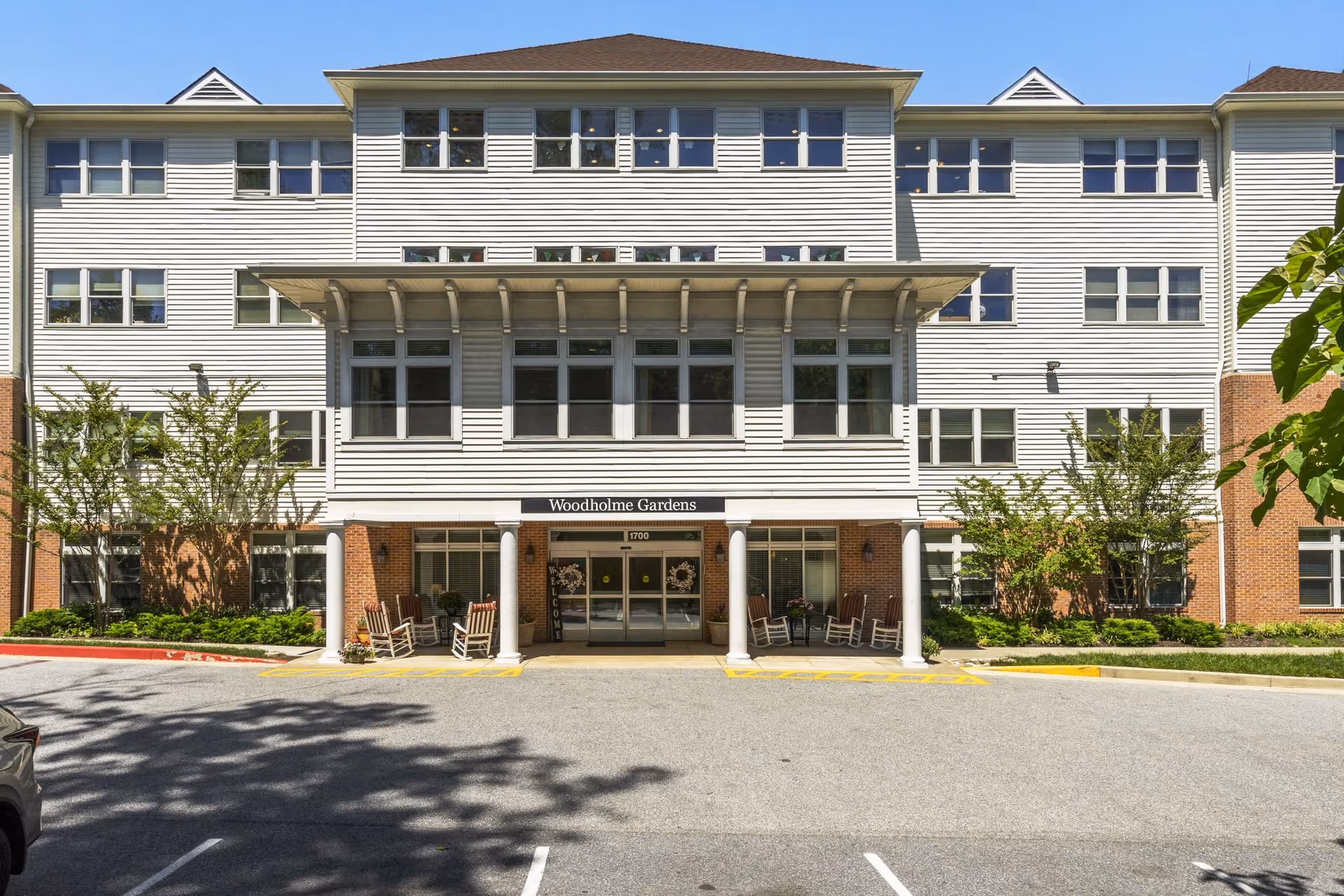 Front exterior view of Woodholme Gardens, a multi-story building with white siding and brick accents. The entrance features a covered porch supported by white columns and several rocking chairs on either side. There are multiple windows on each floor and some greenery around the building.