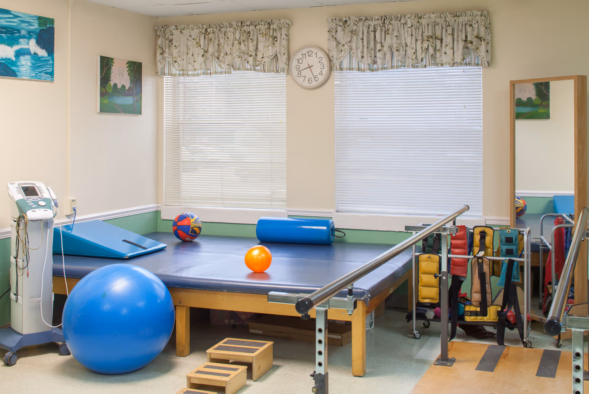 A physical therapy room with exercise equipment including a large blue exercise ball, a padded table with a blue mat, a small orange ball, a colorful basketball, parallel bars for walking support, and various therapy straps hanging on a rack. The room has two windows with floral valances, a clock on the wall, and framed artwork.