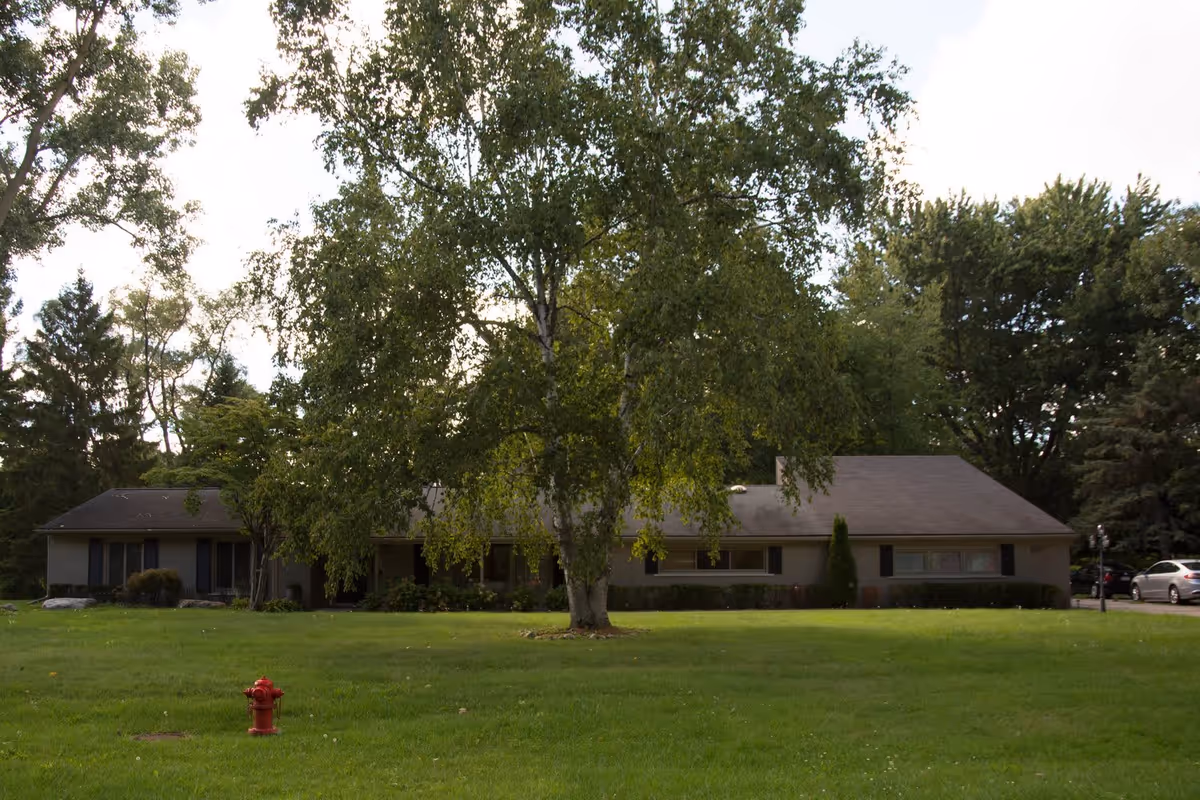 A single-story building partially obscured by a large tree in the front yard, with a well-maintained green lawn and a red fire hydrant in the foreground. Several cars are parked on the right side near the building, and the area is surrounded by tall trees.