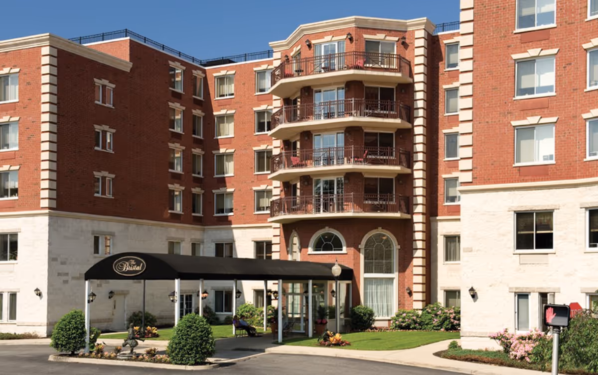 Exterior view of The Bristal Assisted Living at Westbury, showing a multi-story brick building with balconies, a covered entrance with a black canopy displaying the name 'The Bristal', and landscaped greenery around the entrance.