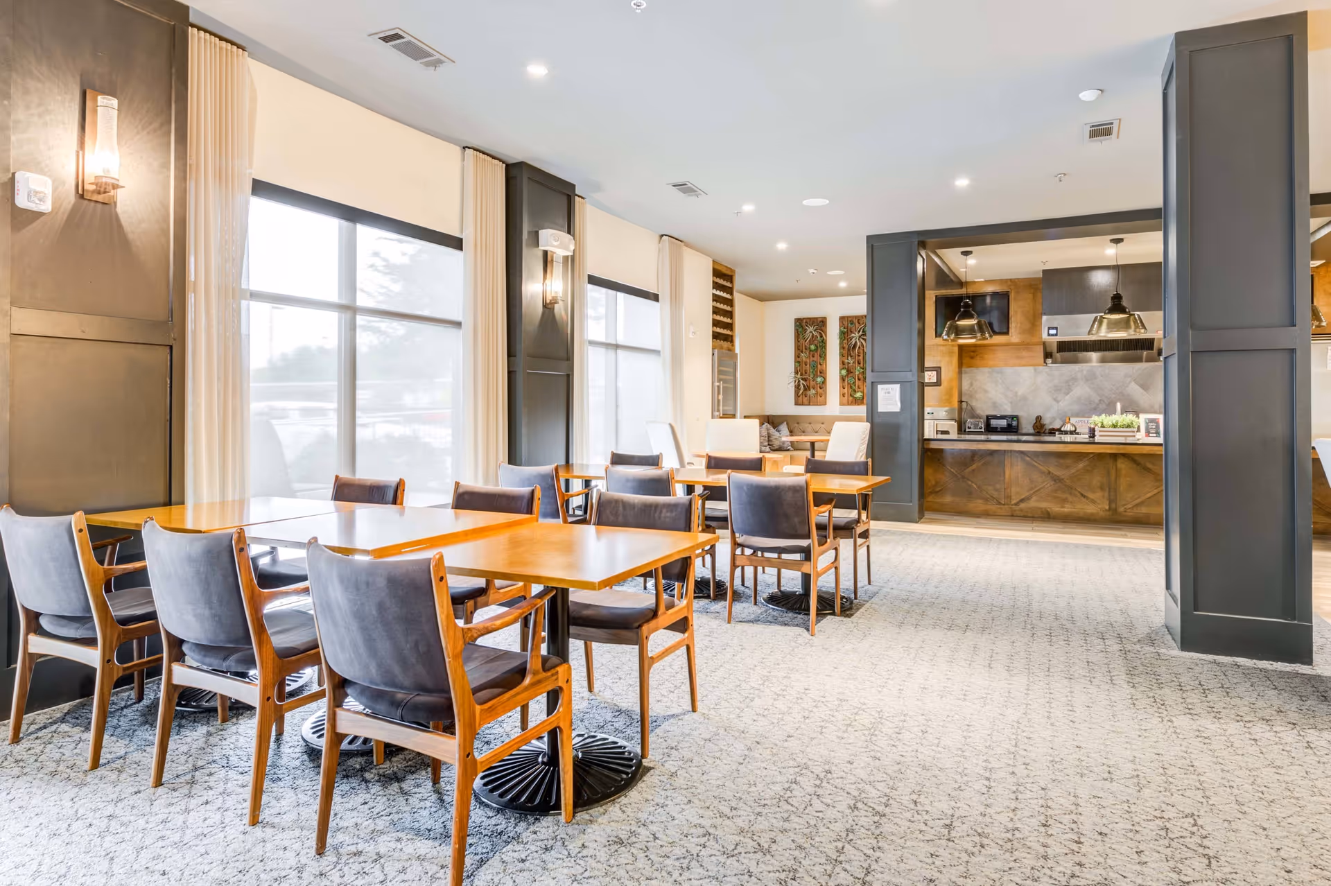 A bright dining area with multiple wooden tables and cushioned chairs arranged neatly on a patterned carpet. Large windows with beige curtains allow natural light to fill the room. In the background, there is a kitchen area with pendant lights, appliances, and wooden cabinetry. The walls are decorated with artwork and sconces.