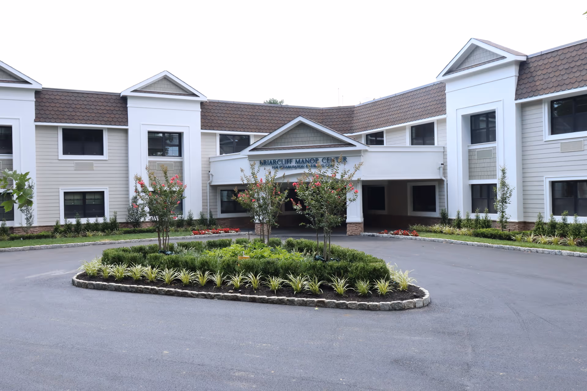 Front exterior view of Briarcliff Manor Center For Rehabilitation And Nursing, showing a two-story building with white and beige siding, multiple windows, a covered entrance, and a circular driveway with landscaped greenery and small trees in the center.