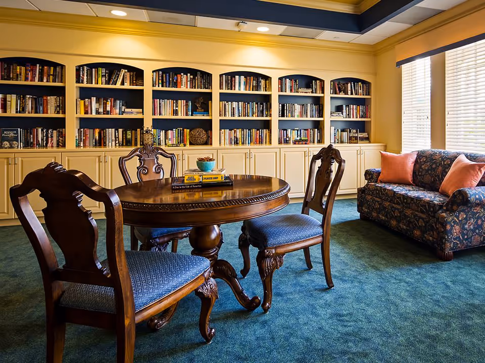 A cozy reading room with a round wooden table surrounded by three ornate wooden chairs with blue upholstered seats. Behind the table is a large built-in bookshelf filled with books and decorative items. To the right, there is a floral-patterned sofa with two pink cushions, next to a window with white blinds allowing natural light to enter. The room has a green carpet and warm yellow walls.