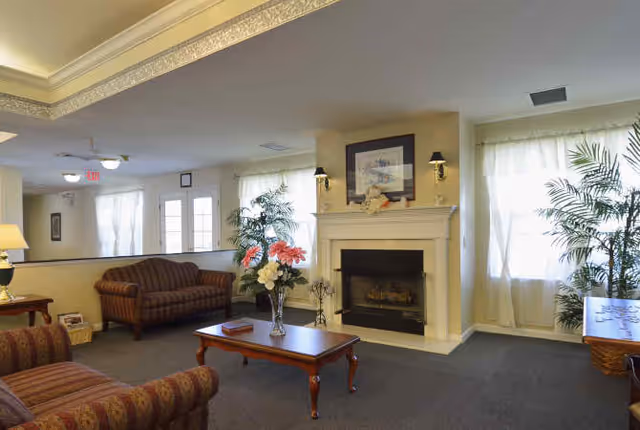A cozy living room area with two patterned armchairs and a matching sofa, a wooden coffee table with a vase of flowers, a white fireplace with a framed picture above it, and large windows with sheer curtains letting in natural light. There are also two potted plants near the windows and a side table with a lamp.