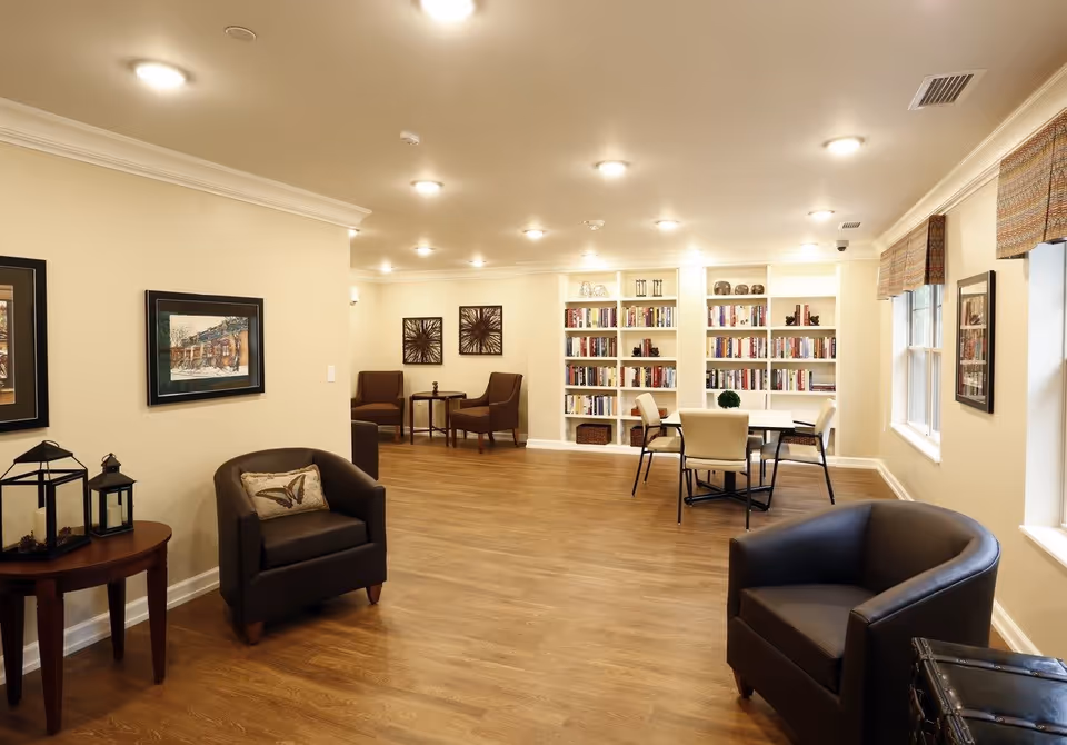 A well-lit common area with wooden flooring featuring two dark leather armchairs, a small round wooden table with decorative lanterns, a seating area with two brown chairs and a small table, a dining table with four chairs, and built-in bookshelves filled with books and decorative items. The walls are light-colored with framed artwork and windows with patterned valances.