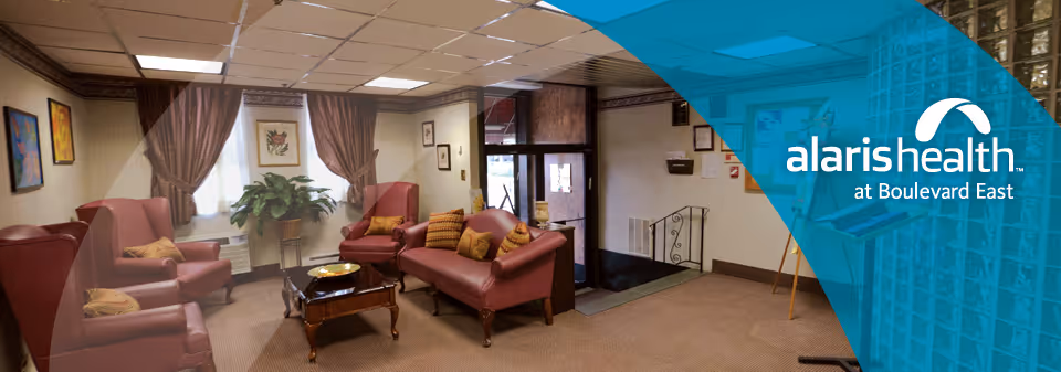 A cozy living room area with several maroon upholstered chairs and a sofa arranged around a wooden coffee table. The room has beige walls adorned with framed artwork and brown curtains on the windows. There is a large green plant near the window and a glass door leading outside.
