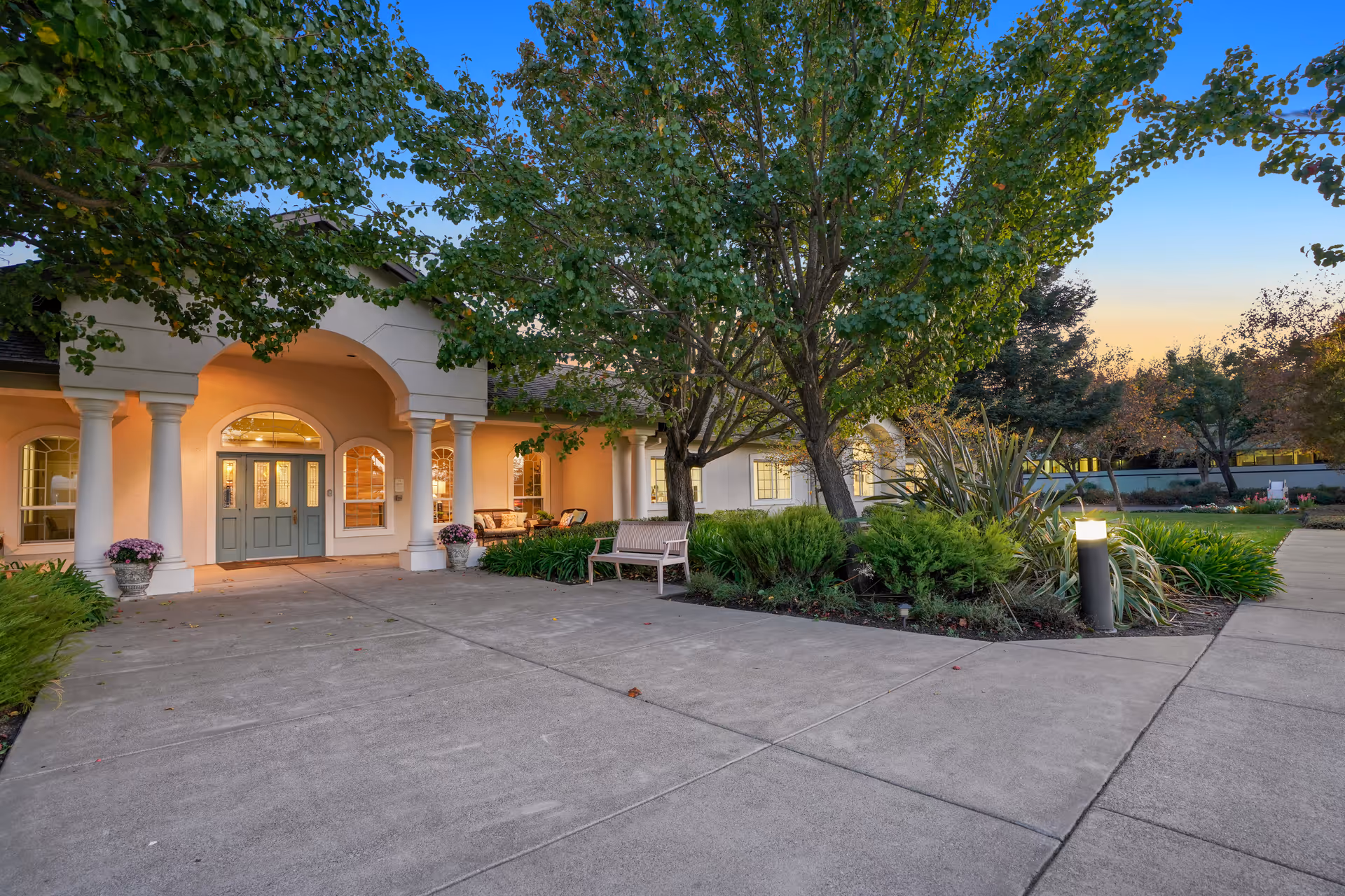 Exterior view of MuirWoods Memory Care facility at dusk, showing a wide concrete driveway leading to a covered entrance with columns and double doors. There are trees, shrubs, and a bench near the entrance, with warm interior lights visible through the windows.