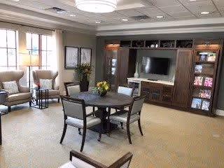 A well-lit living room with a round wooden table and four chairs in the center. The room features a large built-in entertainment center with a flat-screen TV and shelves displaying books and decorative items. Two armchairs are positioned near large windows with white curtains, and framed artwork hangs on the wall. A ceiling light fixture provides additional illumination.