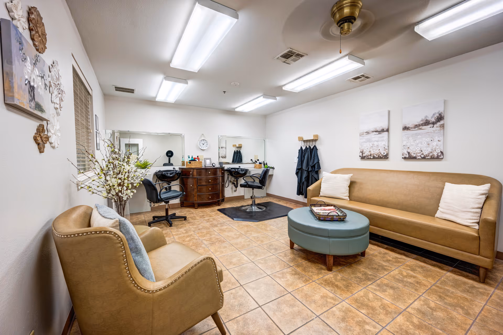 A well-lit room with two salon chairs in front of mirrors and sinks, a brown leather sofa with two white pillows, a matching armchair with a gray throw blanket, a round blue ottoman with a basket on top, wall art, and a coat rack with black capes hanging.