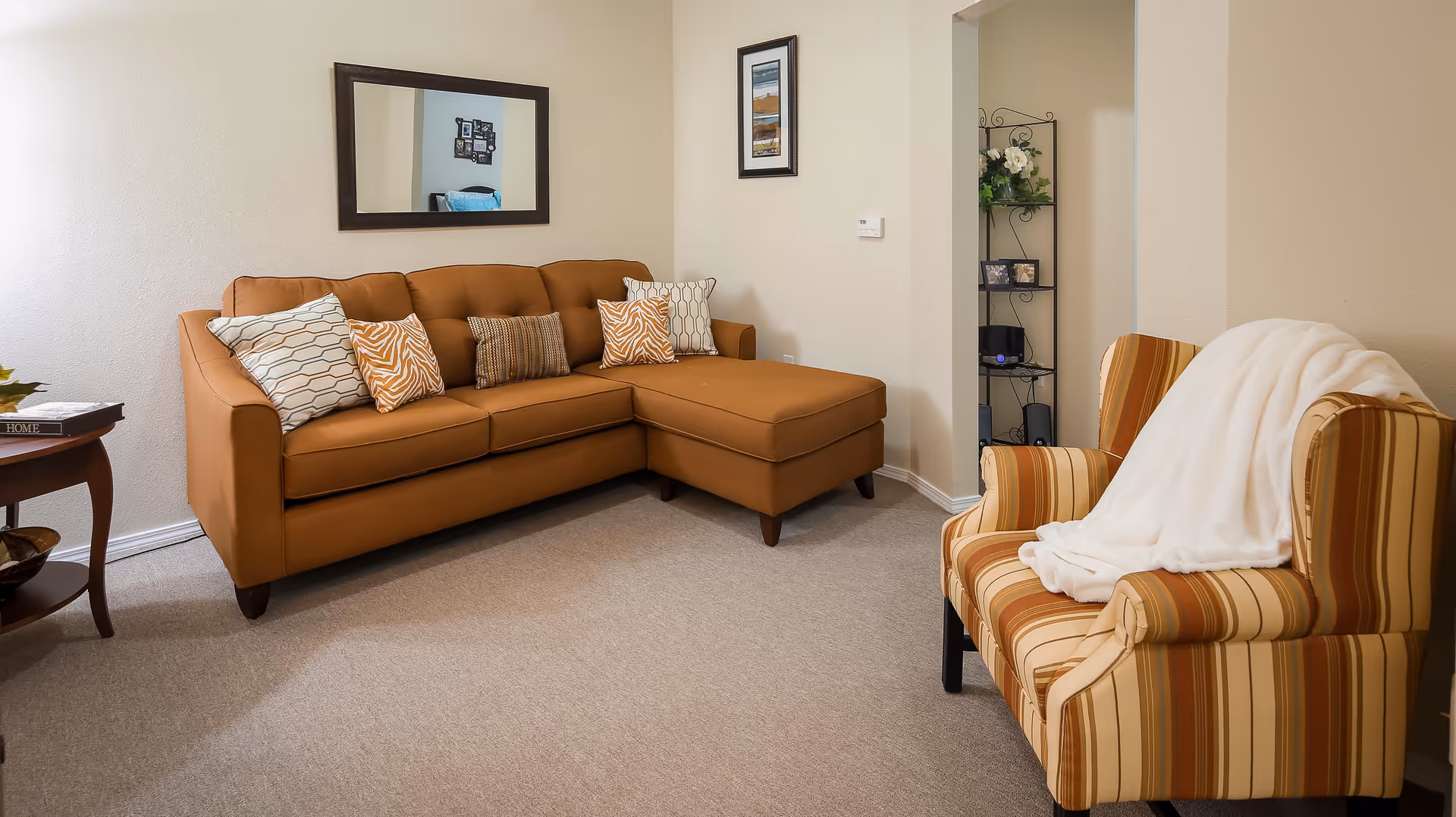 A tidy living room with a tan sectional sofa, patterned throw pillows, a striped armchair draped with a white throw, a wall mirror and a side table.