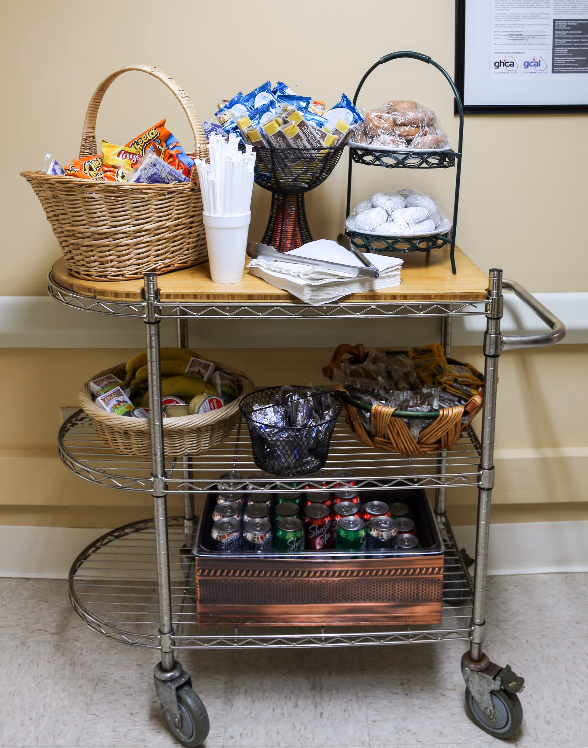 A metal rolling cart with three shelves holding various snacks and drinks. The top shelf has a wicker basket with chips, a cup with white plastic straws, a bowl with granola bars, a two-tiered stand with wrapped pastries, and a stack of napkins with tongs. The middle shelf contains a basket with bananas and juice boxes, a small black basket with wrapped items, and another basket with assorted snacks. The bottom shelf holds a tray filled with assorted canned sodas. The cart is positioned against a beige wall with a framed document partially visible on the right.