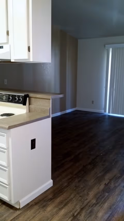 Interior view of a senior living apartment showing a kitchen area with white cabinets, a stove, and a countertop. The kitchen opens into a room with dark wood flooring and vertical blinds covering a sliding glass door.