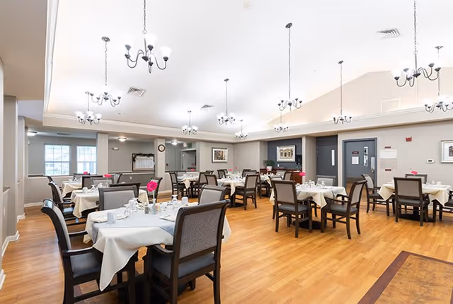 A spacious dining room with multiple tables covered in white tablecloths, each set with cups, plates, and pink flower centerpieces. The room has wooden flooring, beige walls, and several chandeliers hanging from the ceiling providing bright lighting. There are framed pictures on the walls and large windows letting in natural light.
