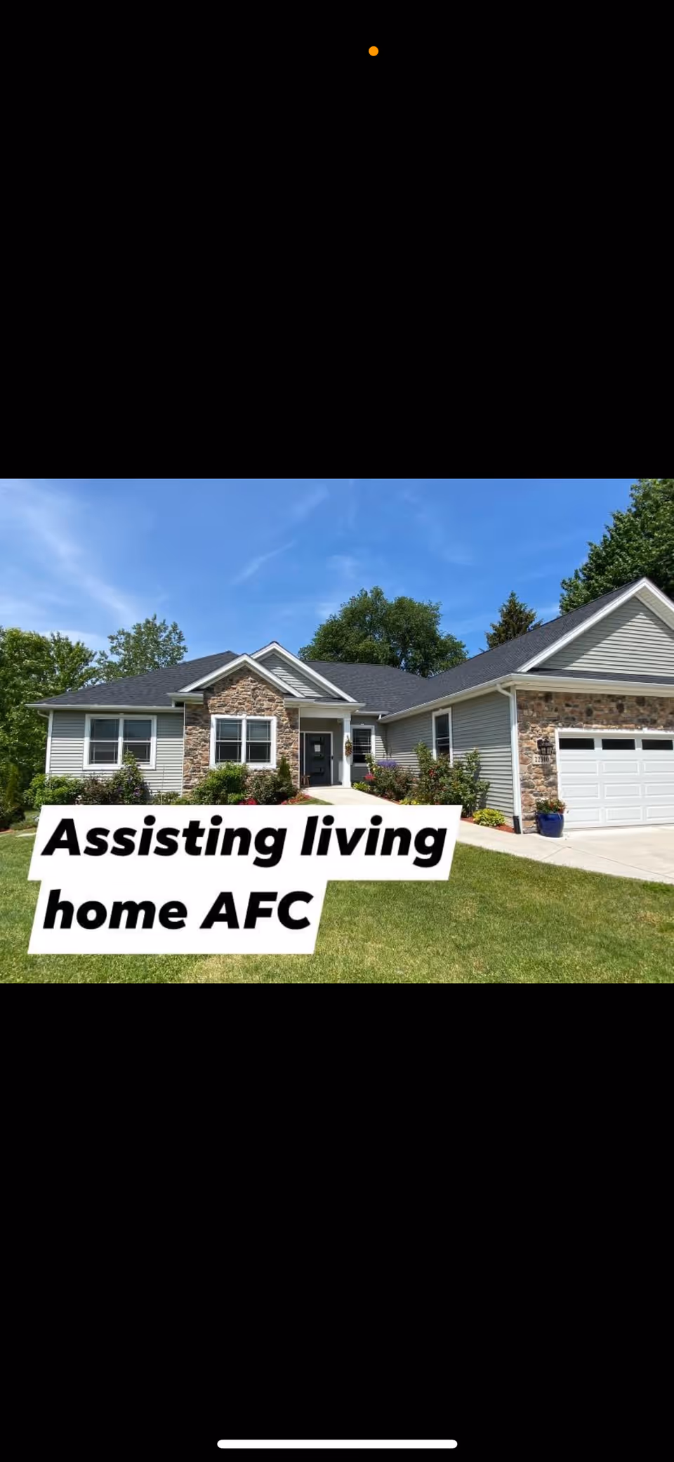 Single-story assisted living home with gray siding and stone accents, a well-maintained lawn, and a driveway leading to a garage under a blue sky.