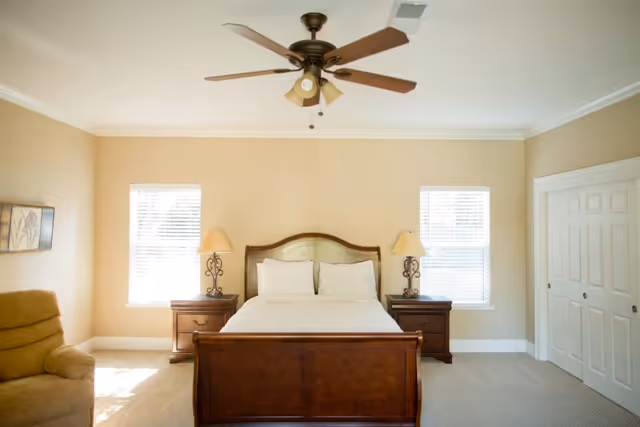 Bright bedroom with a wooden bed centered between two windows, matching nightstands and lamps, a ceiling fan, and a recliner.
