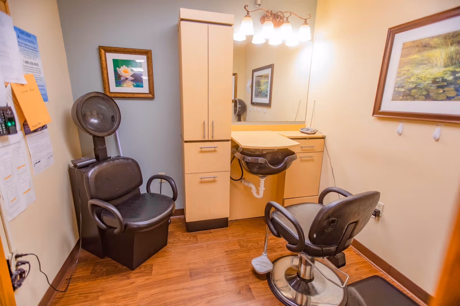 A small salon room with two black salon chairs, one positioned in front of a hair washing sink attached to a wooden cabinet with drawers. The room has wooden flooring, a large mirror above the sink, framed floral artwork on the walls, and some papers pinned on the left wall.