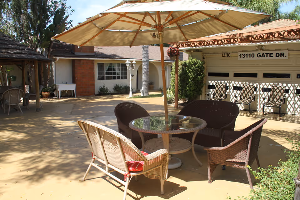 Outdoor patio area with a round glass table and wicker chairs under a large beige umbrella. The patio is paved and surrounded by greenery, with a building in the background displaying the address 13110 Gate Dr.