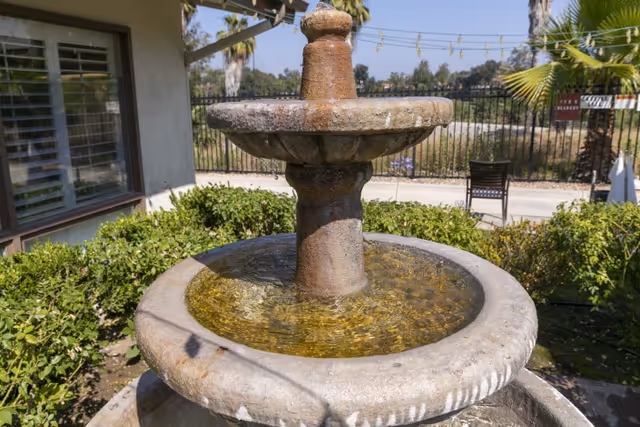 Stone tiered water fountain in a landscaped outdoor courtyard with shrubs, a patio chair, and a building in the background.