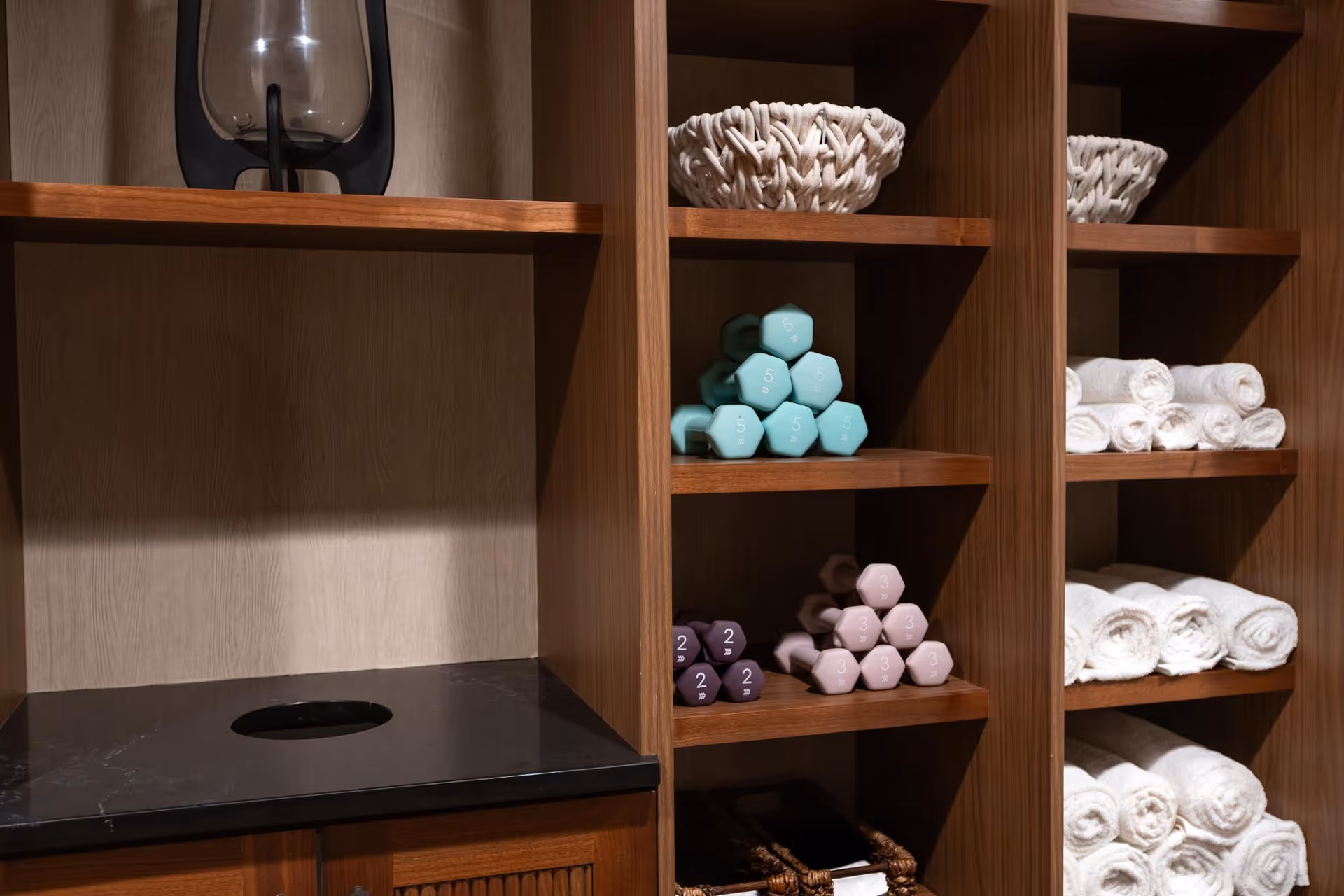 Wooden shelves holding rolled white towels, pastel-colored dumbbells stacked in pyramids, a woven basket, and a glass water dispenser on a black countertop.