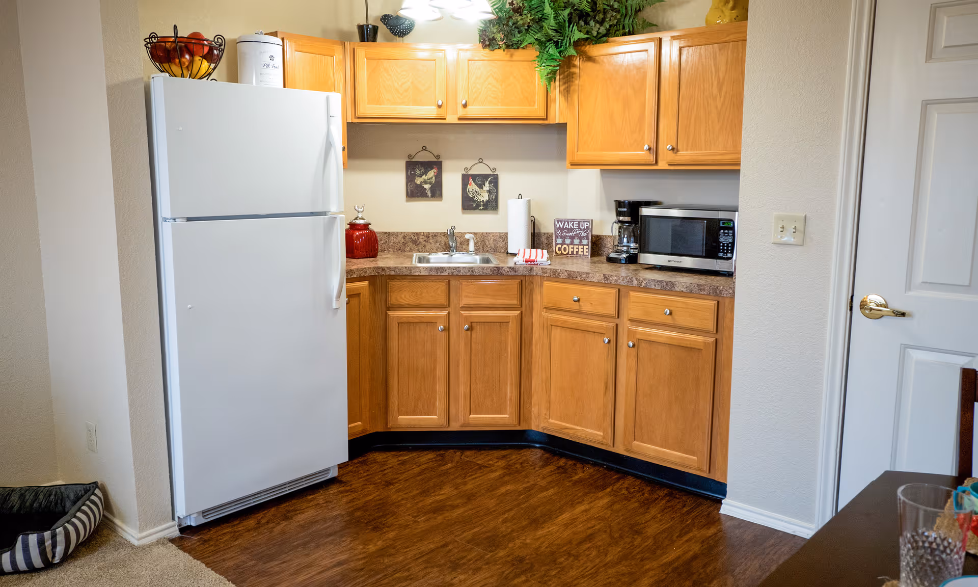 A small kitchen area with wooden cabinets, a white refrigerator, a stainless steel sink, a microwave, a coffee maker, and decorative items including two rooster pictures on the wall and a sign that says 'Wake up & smell the coffee'. There is a fruit basket on top of the refrigerator and some greenery above the cabinets.