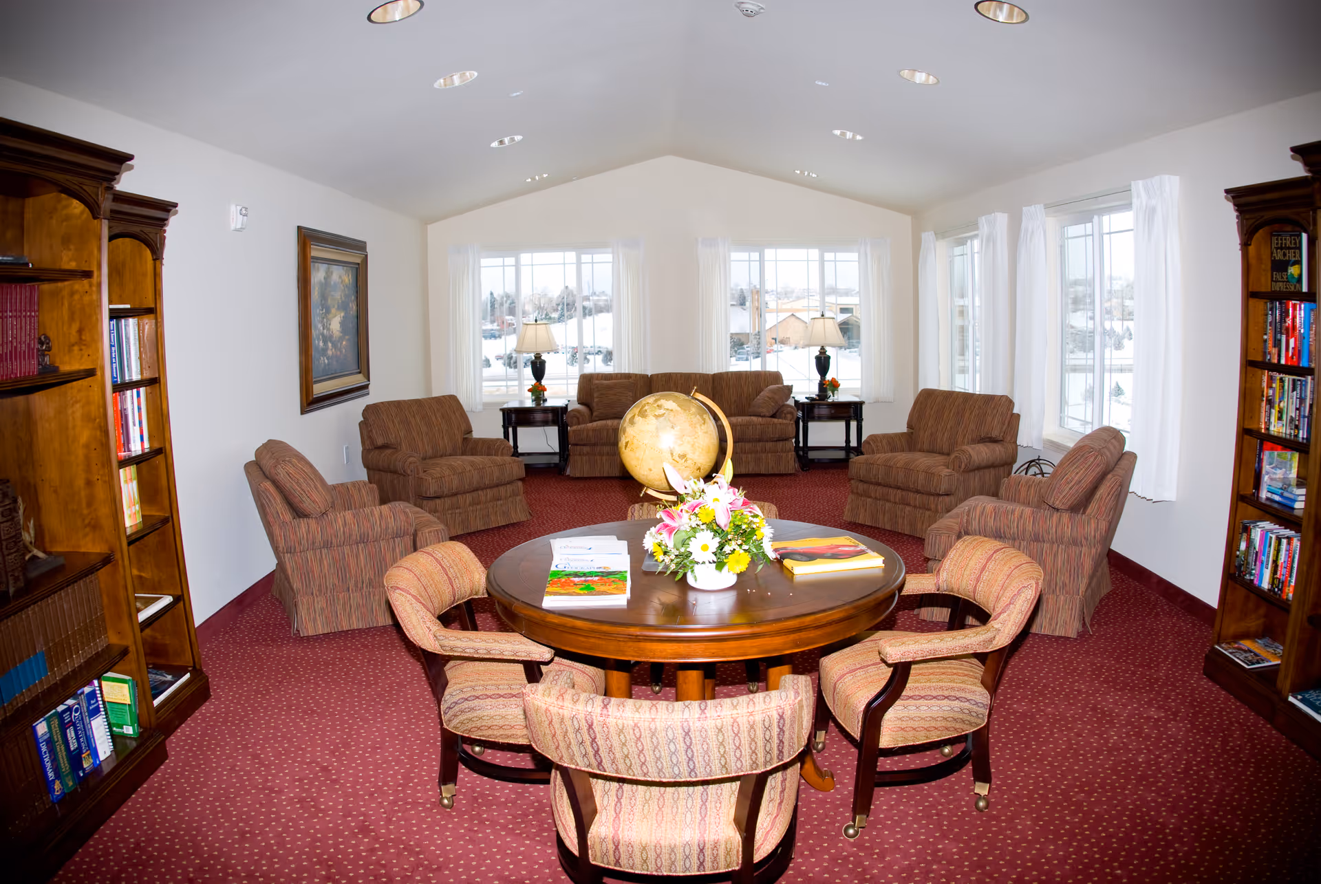 A cozy living room with a round wooden table in the center surrounded by four upholstered chairs. On the table, there is a globe, a flower arrangement, and some books. Around the room, there are six cushioned armchairs arranged in a semi-circle facing the table. The room has large windows with white curtains letting in natural light, two wooden bookshelves filled with books, and a painting on one wall. The carpet is red with a subtle pattern, and the ceiling is vaulted with recessed lighting.