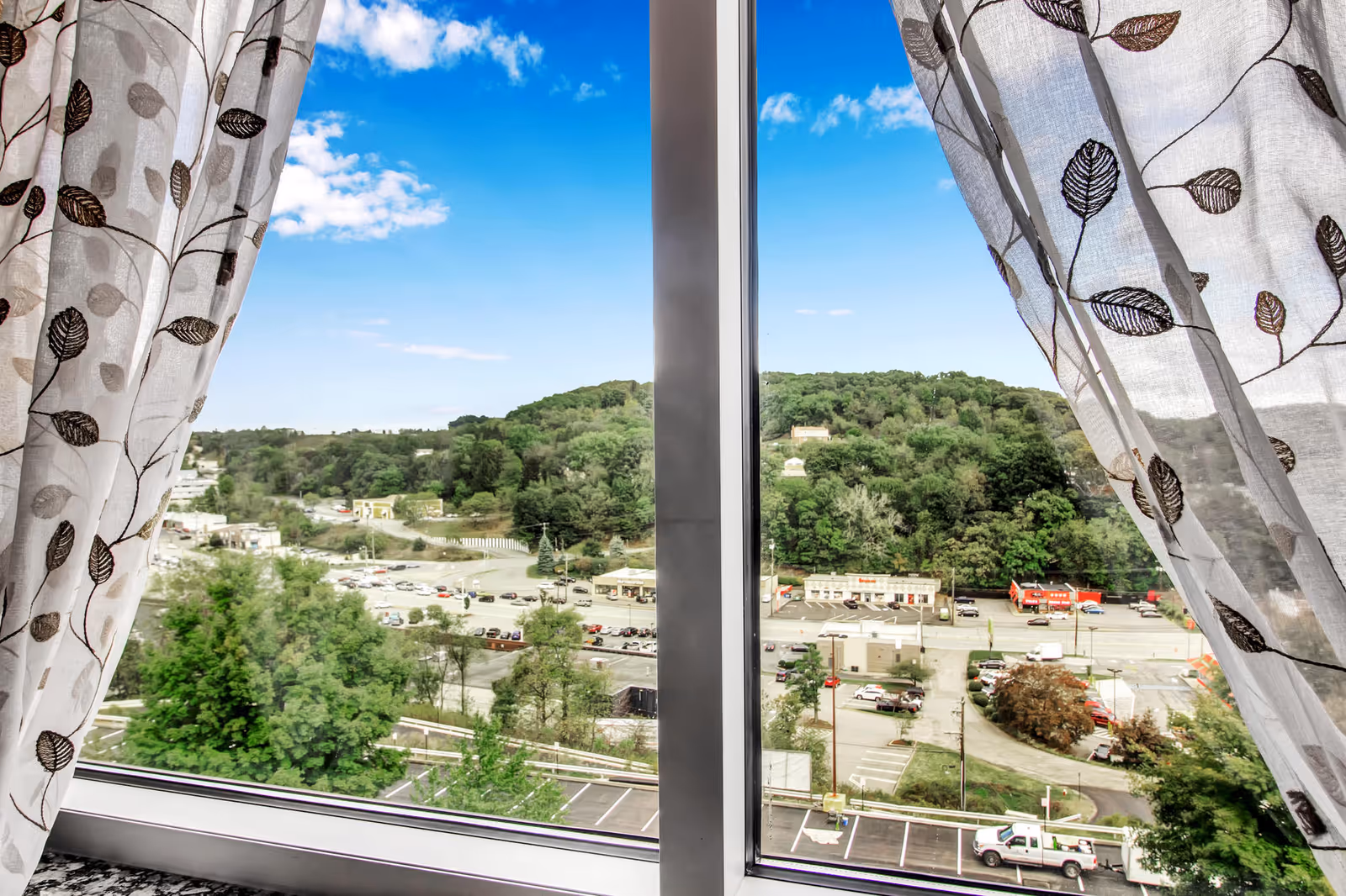 View through a window framed by white curtains with a leaf pattern, showing a parking lot, trees, buildings, and a green hill under a blue sky with some clouds.