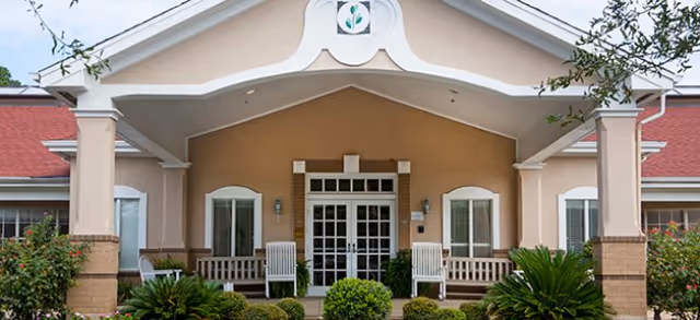 Front exterior view of a building with a covered entrance, two white rocking chairs on either side of the door, and well-maintained shrubs and plants in front.