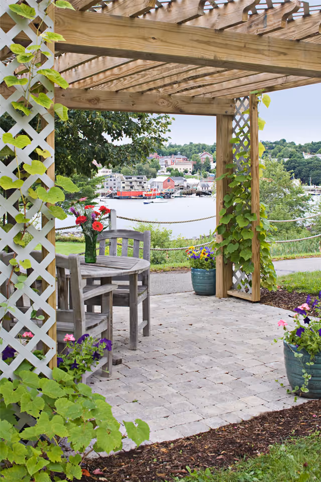 Outdoor patio area with a wooden pergola covered in climbing plants, a round wooden table with two chairs, and flower pots with colorful flowers. In the background, there is a view of a waterfront with boats and buildings across the water.