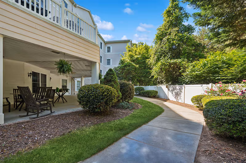 Outdoor patio area with wooden rocking chairs and tables under a covered porch next to a curved concrete walkway surrounded by green bushes, trees, and a white fence on a sunny day.